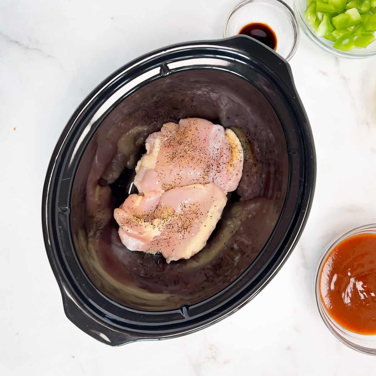 Two seasoned raw chicken thighs in a slow cooker, with small bowls of sauce and chopped green peppers nearby, demonstrating slow cooker chicken tips.