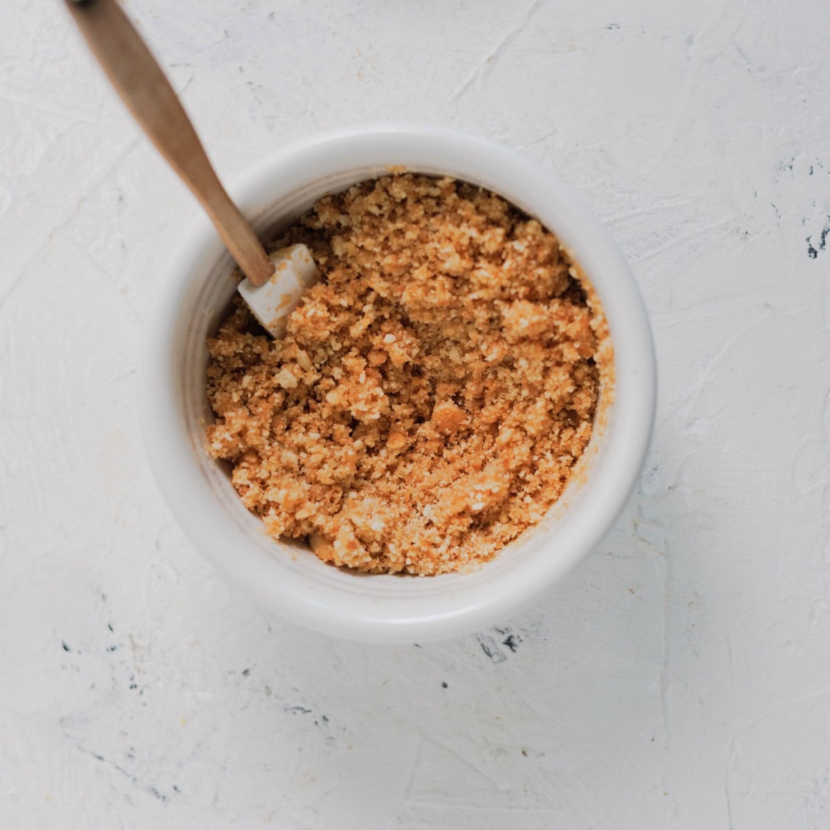 A white bowl filled with golden-brown biscuit crumbs for a no-bake cheesecake, with a wooden spatula resting inside.