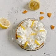 A lemon cream pie topped with whipped cream and lemon slices, surrounded by two lemon halves and cookie crumbs on a white surface.