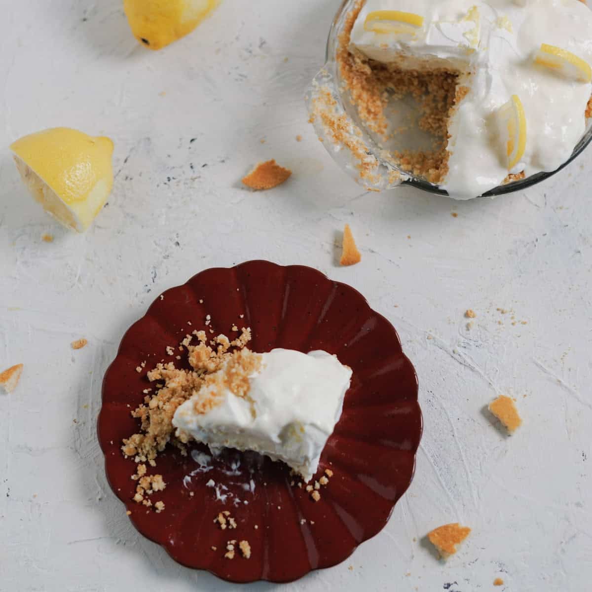 A slice of mini lemon cheesecake on a dark red scalloped plate, with a glass pie dish and lemons nearby.