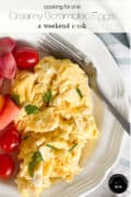 A plate of creamy scrambled eggs garnished with chives, served with cherry tomatoes and melon slices. Text at the top reads, cooking for one, Creamy Scrambled Eggs, a weekend cook.
