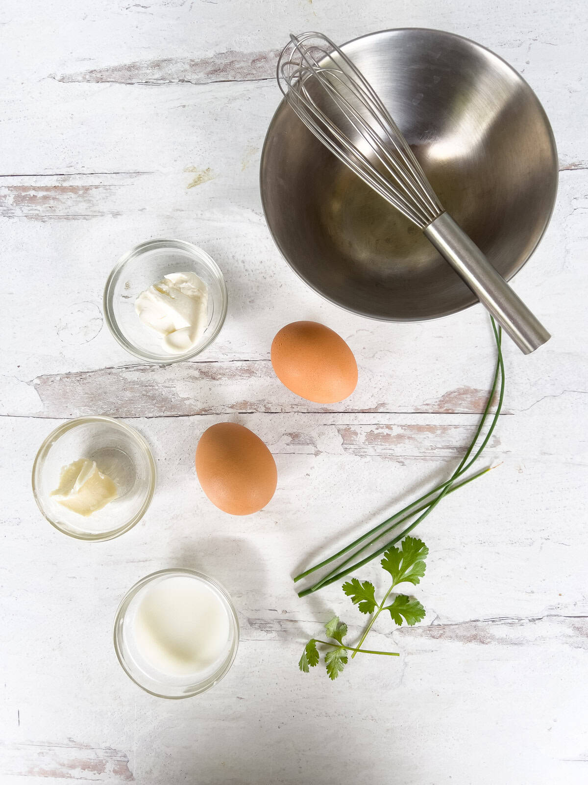 A metal mixing bowl with a whisk, two whole eggs, and small glass bowls containing butter, cream cheese, and milk, alongside sprigs of chives and cilantro.