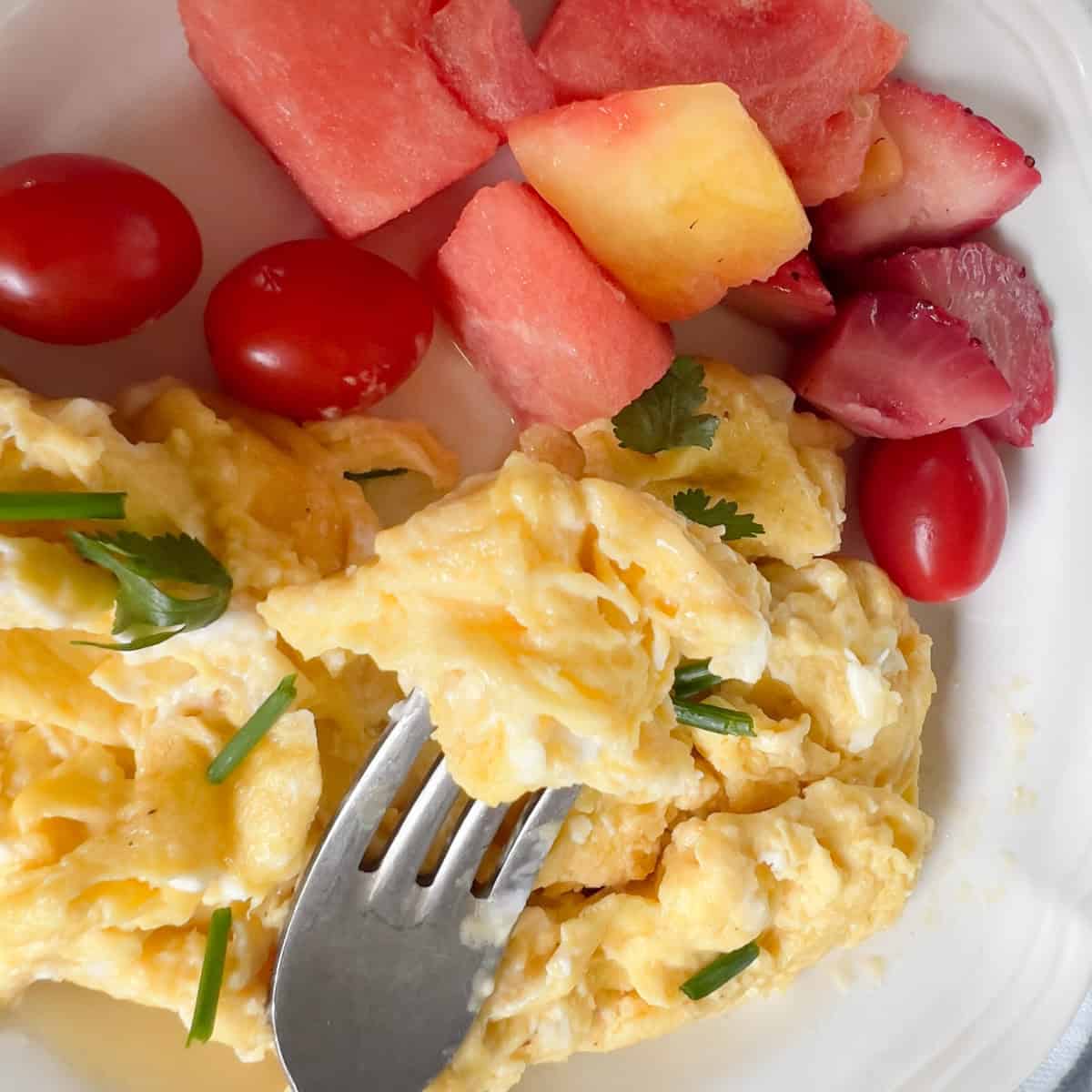 Scrambled eggs garnished with herbs on a plate with cherry tomatoes, watermelon, and strawberries, with a fork lifting a piece of egg.