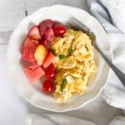 Scrambled eggs garnished with herbs served with a side of watermelon, strawberries, and cherry tomatoes on a white plate with a fork.